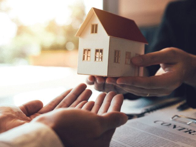 Duas pessoas de terno segurando um modelo de casa em miniatura, representando uma transação imobiliária ou a conclusão de um processo legal de adjudicação.