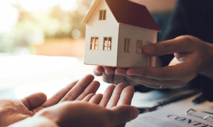 Duas pessoas de terno segurando um modelo de casa em miniatura, representando uma transação imobiliária ou a conclusão de um processo legal de adjudicação.